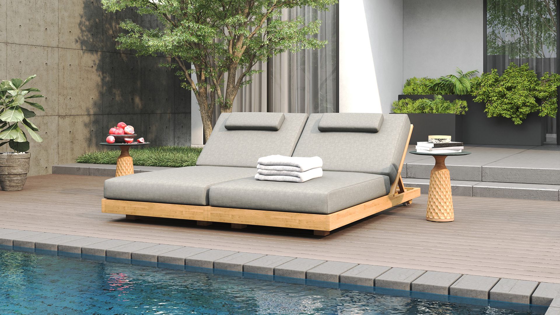 A double teak chaise lounger with soft grey cushions and two side tables on a deck next to a pool.