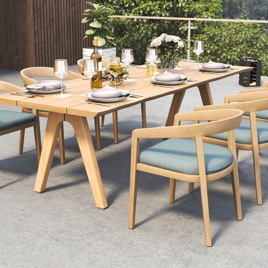 Outdoor dining set on a patio, with a long table and six wooden dining chairs with blue cushions.